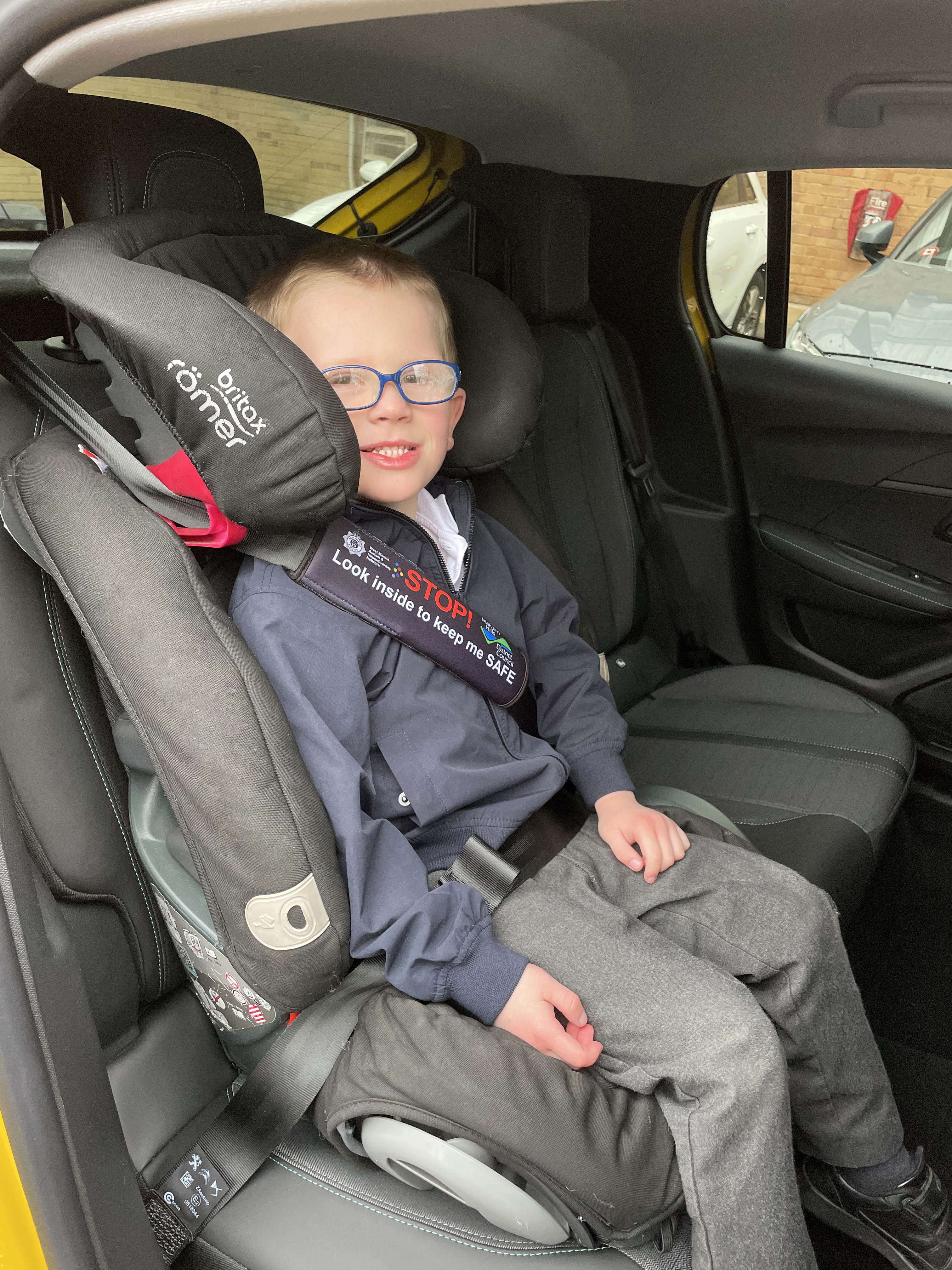 A little boy sitting in a car seat with a seatbelt around him. The seatbelt has a cover that says 'Look inside to keep me safe'.